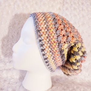 May include: A crocheted beanie hat in shades of gray, peach, and yellow. The hat has a textured stitch pattern and is shown on a white mannequin head.