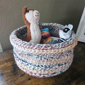May include: A crocheted basket with a white, gray, blue, red, and yellow striped pattern. The basket is filled with a brown and white stuffed llama, a black and white stuffed animal, and a small bag of chips.