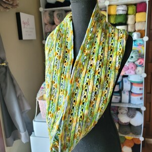 May include: A crocheted infinity scarf in shades of green, blue, and yellow. The scarf is displayed on a black mannequin.