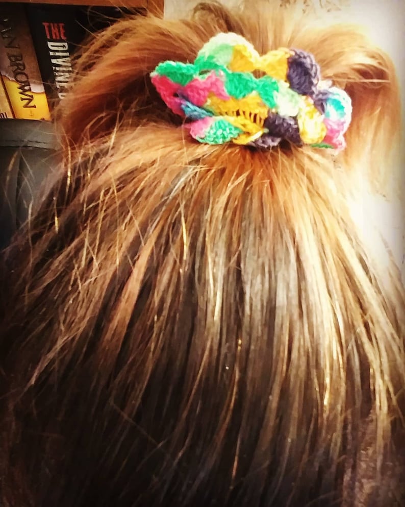 Peut inclure: Un chouchou en crochet en forme de fleur color&eacute; dans des tons de rose, vert, jaune et violet. Le chouchou est nou&eacute; autour d'une queue de cheval de cheveux bruns.