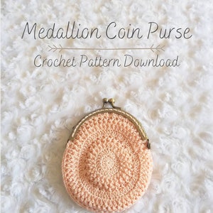 Peut inclure: Une bourse &agrave; monnaie en crochet avec un motif de m&eacute;daillon de couleur p&ecirc;che. La bourse a un fermoir en laiton et est pos&eacute;e sur une surface blanche et moelleuse. Le texte "Medallion Coin Purse Crochet Pattern Download" est au-dessus de la bourse.