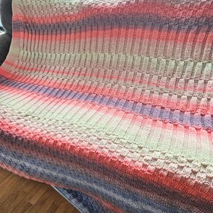May include: A knitted blanket with a gradient color scheme, transitioning from light green to pink, purple, and blue. The blanket features a textured pattern with alternating ribbed and square sections, creating a visually appealing design. The blanket is draped over a sofa.