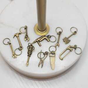 May include: A set of ten gold-toned metal keychains featuring various tools, including a hammer, saw, wrench, screwdriver, and drill. The keychains are arranged on a white surface.