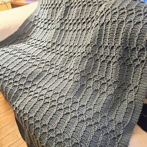 May include: A gray crocheted blanket with a diamond pattern. The blanket is draped over a surface, showcasing the texture and design. The color is a muted gray, suitable for various home decor styles.