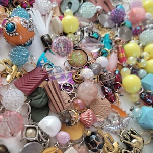 May include: A close-up of a variety of colorful beads, charms, and pendants. The assortment includes glass beads, metal charms, and wooden beads. The colors are bright and vibrant, including pink, yellow, blue, green, and orange.