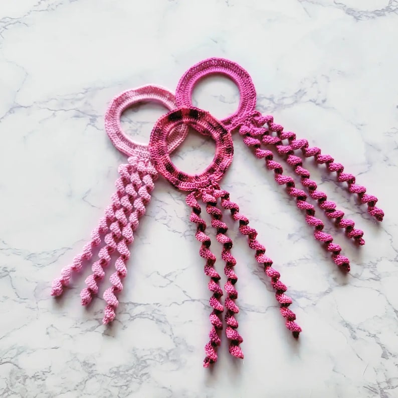 May include: Three pink and purple crocheted hair ties with long, twisted tassels.