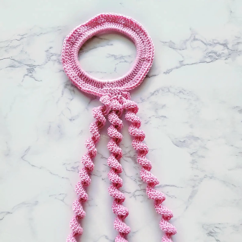 May include: A pink crocheted macrame plant hanger with a circular top and four long, twisted strands.