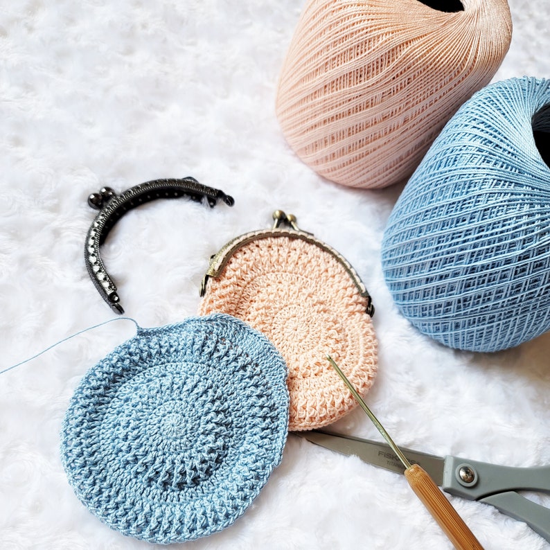 Peut inclure: Deux porte-monnaie en crochet, l'un rose p&acirc;le et l'autre bleu clair, avec un crochet &agrave; crochet et des ciseaux. Les porte-monnaie sont faits avec un point textur&eacute; et ont un fermoir en m&eacute;tal.