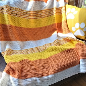 May include: A crocheted throw blanket with stripes of white, orange, and yellow. The blanket is draped over a brown couch with a yellow throw pillow with white paw prints.