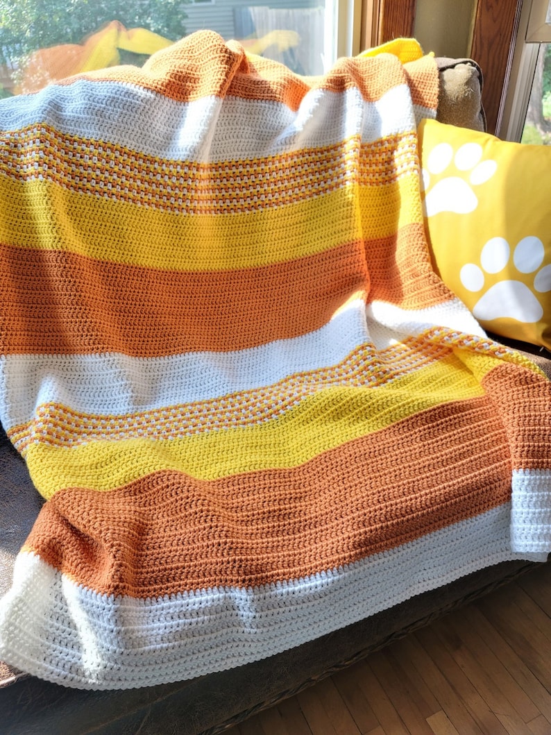 May include: A crocheted throw blanket with stripes of white, orange, and yellow. The blanket is draped over a brown couch with a yellow throw pillow with white paw prints.