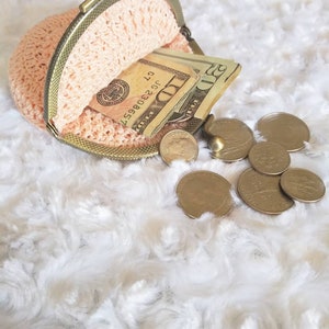 Peut inclure: Une bourse &agrave; monnaie en crochet avec un cadre en laiton est ouverte, r&eacute;v&eacute;lant des billets de dollars am&eacute;ricains et de la monnaie. La bourse est pos&eacute;e sur une surface blanche et moelleuse.