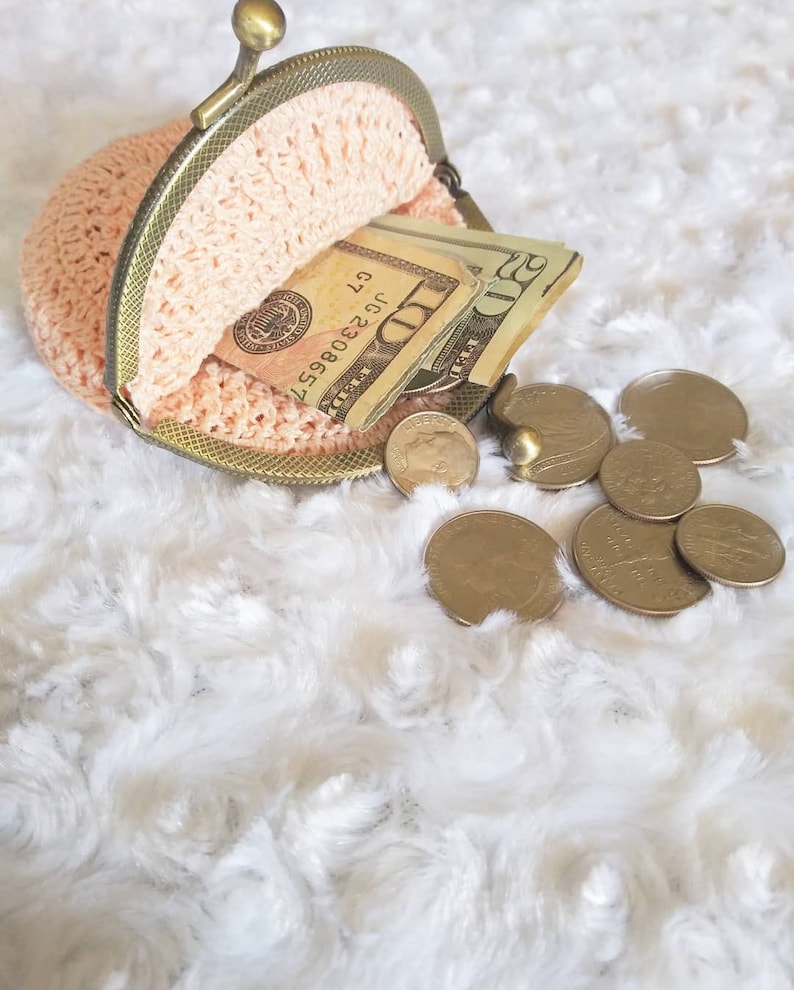 Peut inclure: Une bourse &agrave; monnaie en crochet avec un cadre en laiton est ouverte, r&eacute;v&eacute;lant des billets de dollars am&eacute;ricains et de la monnaie. La bourse est pos&eacute;e sur une surface blanche et moelleuse.