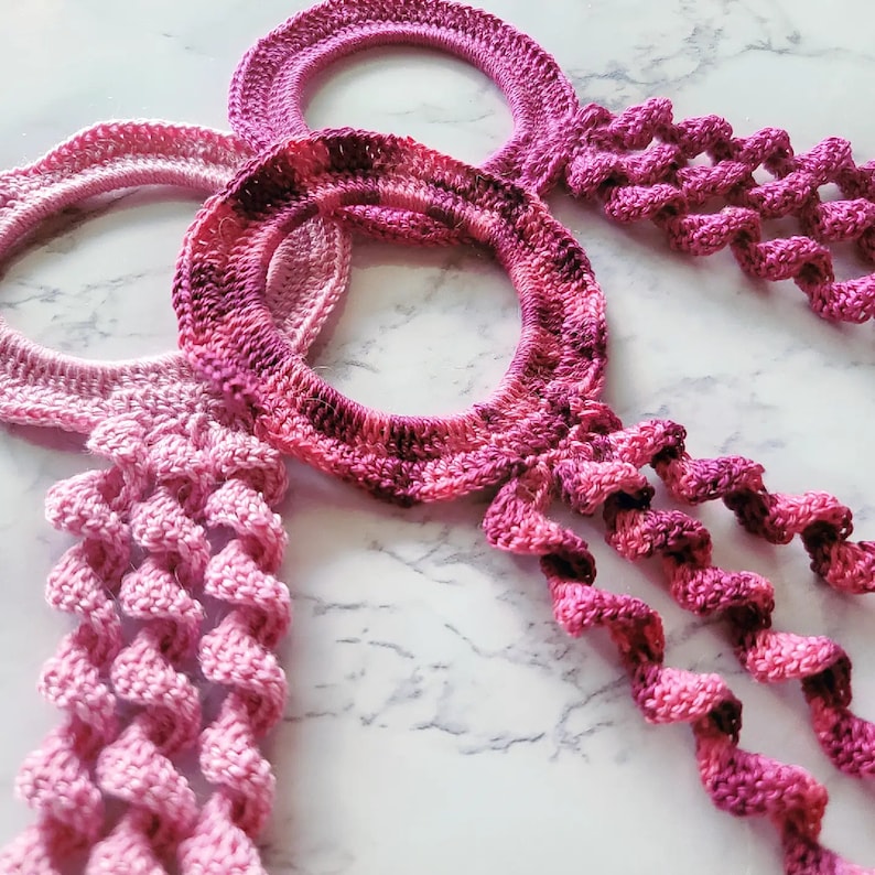 May include: Three crocheted pink and purple hoops with long, twisted tails. The hoops are made with a gradient yarn that transitions from pink to purple.