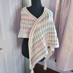 Pode incluir: Um xale de crochê em tons pastel com um padrão quadriculado. O xale apresenta um design de decote em V e é preso com um fecho prateado. Tassels creme pendem dos cantos inferiores. A paleta de cores inclui rosas, verdes e roxos suaves.