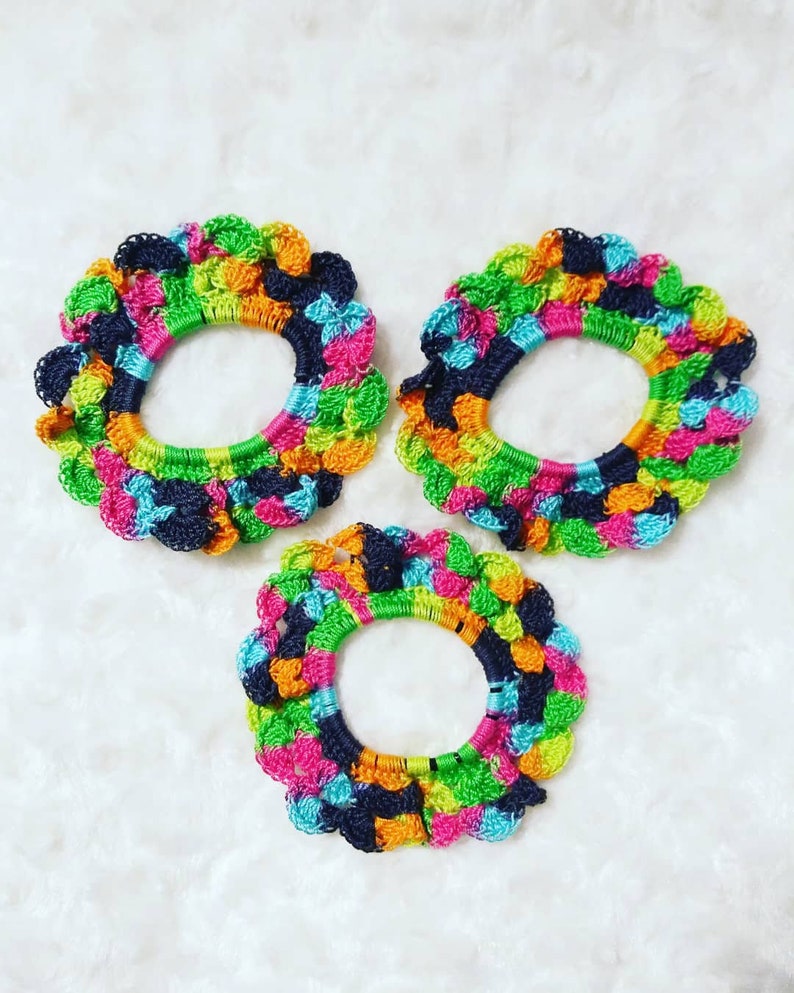 Peut inclure: Trois &eacute;lastiques &agrave; cheveux en crochet color&eacute;s dans des tons de vert, bleu, rose, orange et noir. Les &eacute;lastiques &agrave; cheveux sont dispos&eacute;s en forme de triangle sur une surface blanche et duveteuse.