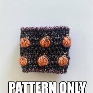 Pode incluir: Aquecedor de chávena de café de crochê com um padrão de listras pretas e brancas e enfeites de abóbora laranja. O texto "PATTERN ONLY" está impresso abaixo do aquecedor.