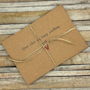 Pode incluir: Um envelope de papel castanho amarrado com barbante e um coração vermelho. O envelope tem o texto "Open when you fancy something sweet" escrito nele.