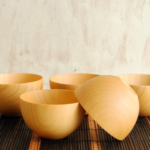 May include: Four wooden bowls, all the same size and shape, are arranged on a brown bamboo mat. The bowls are made of light brown wood and have a smooth, rounded surface.