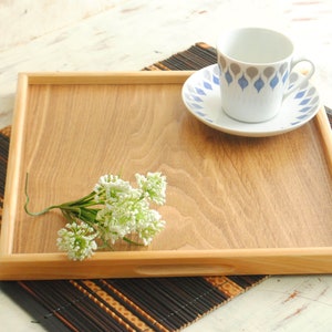 May include: A square wooden serving tray with a light brown wood grain finish and integrated side handles. A white ceramic cup and saucer with a blue and grey teardrop pattern sits on the tray, alongside a small cluster of white flowers.