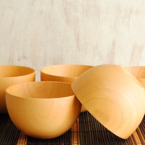 May include: Four wooden bowls of varying sizes are arranged on a brown bamboo mat. The bowls are made of light brown wood and have a smooth, rounded shape.