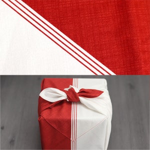 May include: A red and white fabric gift wrap tied with a bow. The fabric has a diagonal design with red and white sections. The red section has three white stripes. The gift wrap is on a gray surface.