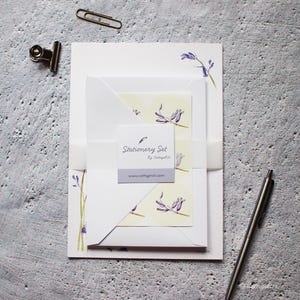 Pode incluir: Um conjunto de papelaria com um design floral com flores roxas sobre um fundo branco. O conjunto inclui papel para escrever cartas, envelopes e um cart&atilde;o com o texto "Stationery Set by CottageGirl" e o endere&ccedil;o do site "www.cottagegirl.com".