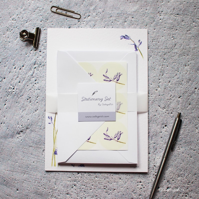Pode incluir: Um conjunto de papelaria com um design floral com flores roxas sobre um fundo branco. O conjunto inclui papel para escrever cartas, envelopes e um cart&atilde;o com o texto "Stationery Set by CottageGirl" e o endere&ccedil;o do site "www.cottagegirl.com".