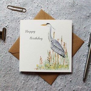 Pode incluir: Um cart&atilde;o branco com o texto "Happy Birthday" e uma ilustra&ccedil;&atilde;o em aquarela de uma gar&ccedil;a-azul em p&eacute; na grama alta.