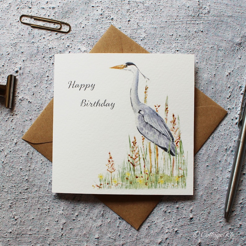 Pode incluir: Um cart&atilde;o branco com o texto "Happy Birthday" e uma ilustra&ccedil;&atilde;o em aquarela de uma gar&ccedil;a-azul em p&eacute; na grama alta.