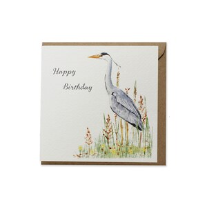 Pode incluir: Uma ilustra&ccedil;&atilde;o em aquarela de uma gar&ccedil;a-azul em p&eacute; na grama alta com o texto "Happy Birthday" em um cart&atilde;o branco.