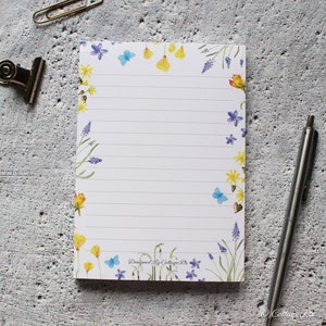 May include: A white notepad with a floral border. The border features blue butterflies, yellow flowers, and purple flowers. The notepad has lined paper and the text "Designed By Cottage Rts" at the bottom.
