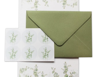 Pode incluir: Conjunto de papelaria com um envelope verde, papel de carta a condizer e autocolantes redondos. O papel e os autocolantes apresentam um design de eucalipto em aquarela em tons de verde e branco. O envelope é de cor verde-oliva lisa.