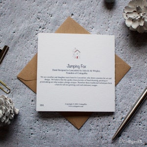 Może przedstawiać: Biała kartka z niebieskim i czerwonym tekstem. Tekst brzmi "Jumping Fox" i "Hand Designed in Lancashire by Julie & Ali Wrigley, Founders of CottageRts". Kartka zawiera r&oacute;wnież małą ilustrację domu z czerwonymi drzwiami.