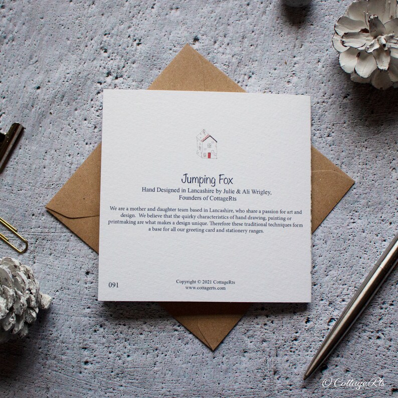 Może przedstawiać: Biała kartka z niebieskim i czerwonym tekstem. Tekst brzmi "Jumping Fox" i "Hand Designed in Lancashire by Julie & Ali Wrigley, Founders of CottageRts". Kartka zawiera r&oacute;wnież małą ilustrację domu z czerwonymi drzwiami.