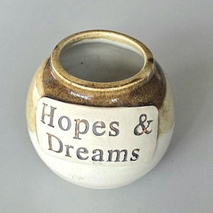 May include: A small, round ceramic jar with a wide opening. The jar is primarily white with a band of brown around the top and a rectangular label that reads "Hopes & Dreams".