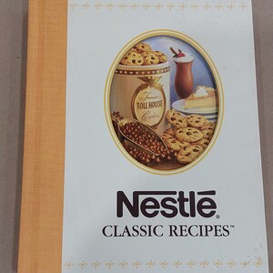 May include: A vintage cookbook with a white cover featuring a colorful illustration of a jar of chocolate chips, cookies, and a glass of milk. The title of the book is "Nestle Classic Recipes".