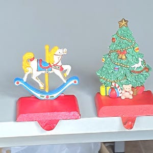 May include: Two Christmas stocking holders. One is a rocking horse with a yellow mane and tail, and the other is a decorated Christmas tree. Both are on red bases with a hook for hanging stockings.
