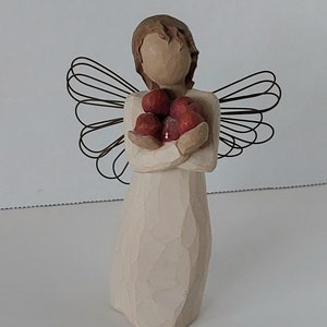 May include: A wooden angel figurine with wire wings holding a bunch of red apples. The angel is wearing a white dress and has a simple, carved face.