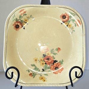 May include: Square, vintage serving plate with a cream-colored background and floral design. The plate features orange and yellow flowers with green leaves. The rim has a rope-like texture and a black border. The plate is displayed on a black metal stand.