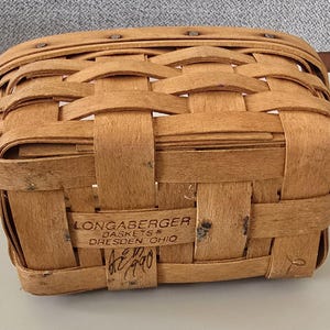May include: A rectangular, woven basket with a light brown hue. It features a tightly woven pattern and two brown leather handles. The basket has the text "Longaberger Baskets Dresden Ohio" and the year "1990" printed on the front.