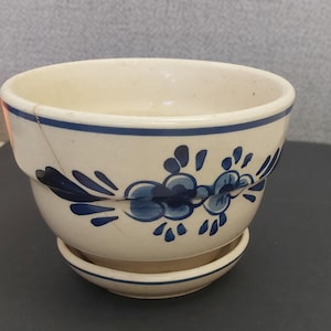 May include: Cream-colored ceramic planter with a matching saucer. The planter features a blue floral design and a blue rim. The planter has a crack on the side. A decorative piece for plants.
