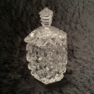 May include: A clear, cut-glass jar with a lid. The jar features a geometric diamond pattern and a decorative finial on the lid. The jar is likely used for storing small items or as a decorative piece.