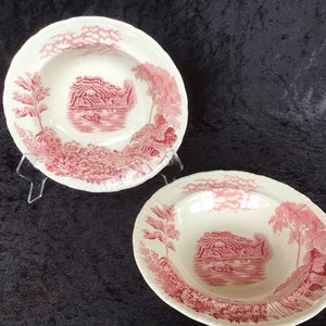 May include: Two white ceramic bowls with a red landscape design. The bowls have a scalloped edge and a detailed illustration of a scene with trees, water, and a building. The bowls are displayed on a clear stand against a dark background.