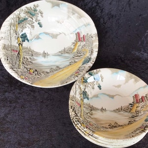 May include: A collection of vintage ceramic bowls, each adorned with a detailed landscape scene. The bowls display a river, trees, and a castle in grey, yellow, and red hues. The bowls are stacked on a dark surface.