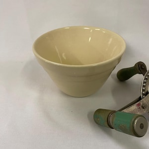 May include: A cream-colored ceramic mixing bowl with a rounded shape and horizontal ridges. A vintage hand-cranked kitchen tool with green handles is partially visible in the background. The bowl is on a white surface.