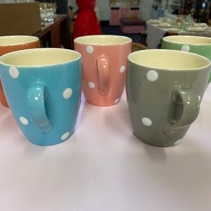 May include: Five ceramic mugs in shades of orange, blue, pink, grey, and green. Each mug has a white polka dot design and a curved handle. The mugs have a cream-coloured interior.