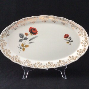 May include: Oval serving platter with a scalloped edge. The white ceramic platter features a red rose and gold floral accents. The rim is decorated with a gold floral pattern. The platter is displayed on a clear plastic stand.