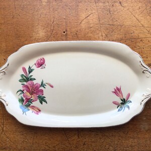 May include: Cream-coloured oval serving platter with a floral design. The platter features pink and purple lilies with green leaves and gold trim along the edges. The edges have a scalloped design.