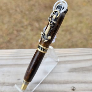 May include: A brown and gold pen with an anchor and chain design. The pen is on a clear acrylic stand.