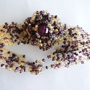 May include: A beaded bracelet featuring a central floral design with a large, faceted, purple gemstone. The bracelet is made of small, clear, gold, and purple beads. The bracelet is approximately 6 inches in length.